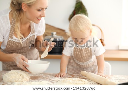 Little girl and her blonde mom in beige aprons  playing and laughing while kneading the dough in kitchen. Homemade pastry for bread, pizza or bake cookies. Family fun and cooking concept