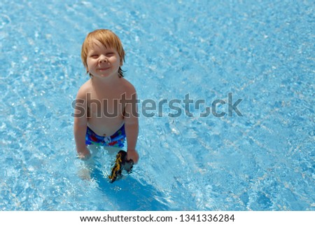 Little relaxed satisfied fair toddler boy in blue shirts standing with goggles in his hand in the warm water of swimming pool in the sunshine