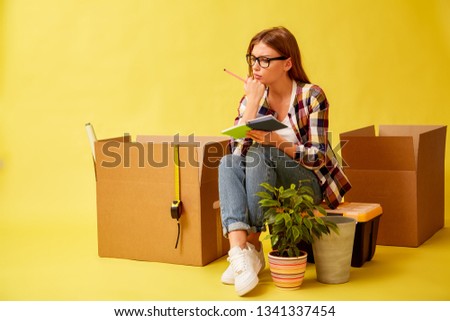Young girl holding a notepad, sitting on a tool box, between boxes for moving. Yellow background.