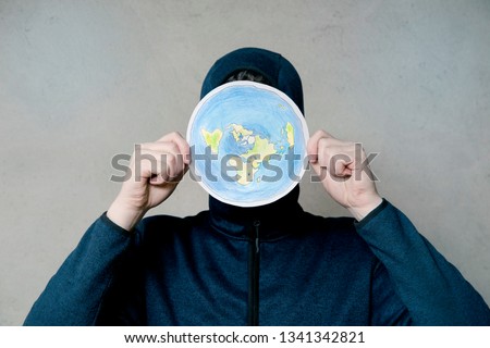 Flat Earther concept. Person who believes that Earth is flat disc. Anonymous hooded Man holding flat Earth model in front of face, copy space. Isolated on gray background, studio shot.