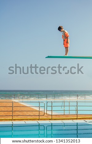 Boy with sleeve floats standing on high spring board and looking at swimming pool below. Boy learning to dive from diving platform at outdoor swimming pool. Royalty-Free Stock Photo #1341352013
