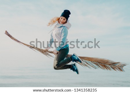 Girl with long hair dressed in pink and grey jacket and grey trousers jumping with palm leaf like a wizard on broomstick and smiling. Blue sky backwards