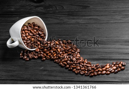 Roasted coffee beans spilling out of a white cup on the wooden table. Natural coffee seeds scattered nicely on grey surface.