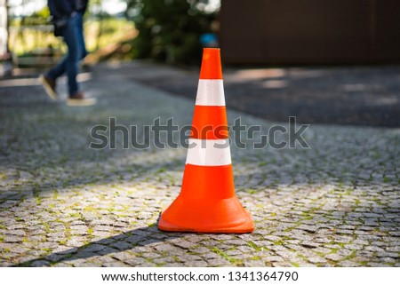Plastic orange parking cone standing in the street on the blurred background of male's legs. Striped road sign for parking in the pavement. Close-up