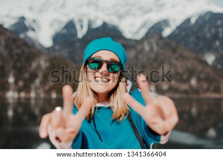 Beautiful girl with short blond hair dressed in green and pink colored hoodie and green hat, wear sunglasses, posing in fron of mirror lake and high mountains with snowy peaks, location lake Ritsa 