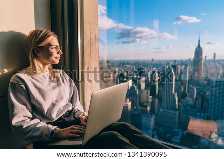 Caucasian female digital nomad sitting with laptop computer on window sill of modern skyscraper building coworking. Young woman freelancer enjoying downtown cityscape from hotel room during vacations Royalty-Free Stock Photo #1341390095
