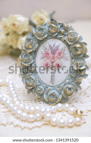 A frame with embroidered flowers, a necklace of pearls and white roses.