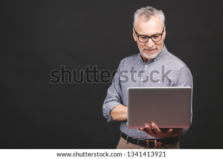 Senior aged bearded old man in eyeglasses holding laptop computer and smiling isolated against black background.