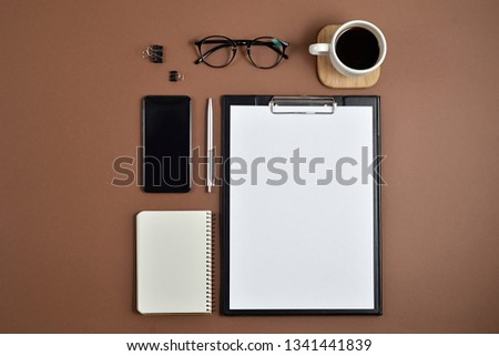 Mockup workspace with blank clip board, office supplies, pen, notepad, smartphone, coffee cup and spectacles on brown background. Flat lay, top view, stylish concept