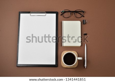 Mock up workspace with blank clip board, office supplies, pen, coffee cup, notepad and eyeglasses on brown background. Flat lay, top view, copy space
