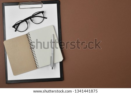 Flat lay, top view office table desk. Workspace with blank clip board, notebook, spectacles, pen on brown background.