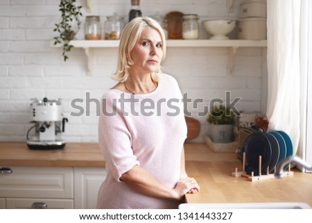 Portrait of attractive gorgeous middle aged European woman with dyed hair standing at kitchen counter in the morning, having serious look, going to make coffee, looking at camera, dressed casually