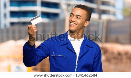 Young afro american worker man holding a credit card and thinking in a construction site