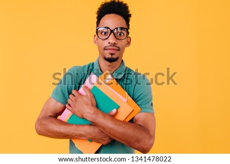 African student in bright t-shirt posing with surprised face expression. Studio photo of black boy in glasses standing on yellow background with books and looking to camera.