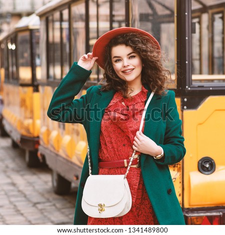 Outdoor portrait of young beautiful happy smiling lady wearing orange hat, snakeskin printed dress, green coat, with white cross body bag, posing in street of European city Royalty-Free Stock Photo #1341489800