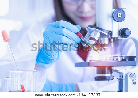 Close up shot of female scientist dropping green liquid from dropper to petri dish on microscope, red liquid in beaker and dropper near her, analysis  experiment to find new experimental results. Royalty-Free Stock Photo #1341576371