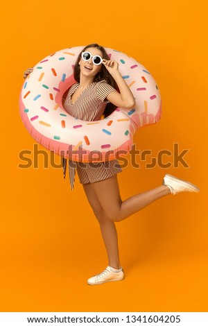 Vertical full length picture of stylish teenage girl in summer dress, sunglasses and sneakers posing with inflatable pink circle, going to have swim in swimming pool during vacations in resort area