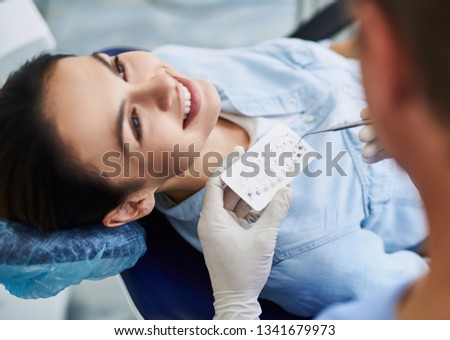 Close up of stomatologist hands in sterile gloves holding sample of bracket system and dental explorer while charming lady lying in dentist chair. Girl is smiling on blurred background Royalty-Free Stock Photo #1341679973