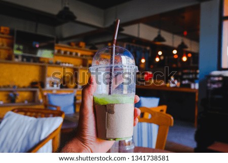 Hand holding green tea plastic cup in coffee shop environment.  Man drink green tea. Hand of man holding Ice tea. Coffee shop environment. Almost all the glasses. Cold Tea Concept