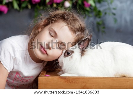 Little girl playing with real rabbit. Child and white bunny on Easter on flower background. Kid kiss pet. Fun and friendship for animals and children.