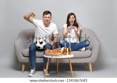 Expressive couple woman man football fans cheer up support favorite team showing thumb down using mobile phone isolated on grey wall background. People emotions sport family leisure lifestyle concept