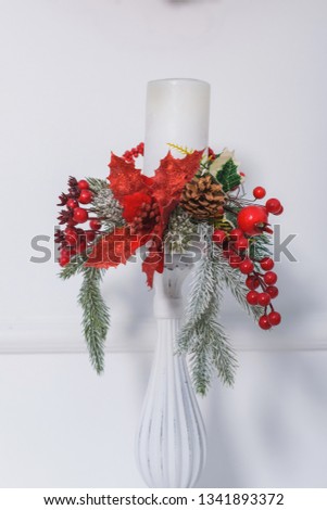 The candle is decorated with pine branches. Red flowers poinsettia. Shelf with candles over the fireplace. Christmas decor. White candlestick.