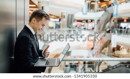Cheerful young  economist  holding laptop and showing thumbs up, happy, dressed in black suit, positive facial emotions, indoor, profile view,  mall background