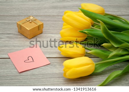 Yellow tulips lie on a gray wooden table next to a sticker on which a heart is drawn. Flowers with a note and a gift for the beloved.