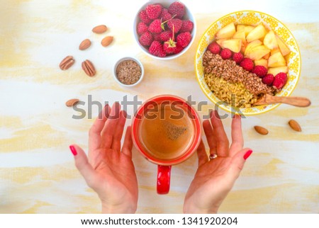 Top view, female hands holding a cup of coffee, a spoon with oatmeal, with honey nuts, raspberries, on a yellow wooden table Good morning - healthy breakfast background