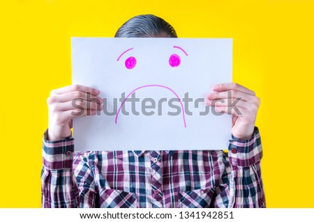 A girl in a plaid shirt on a yellow background holds in her hands a white sheet of paper with a sad face painted