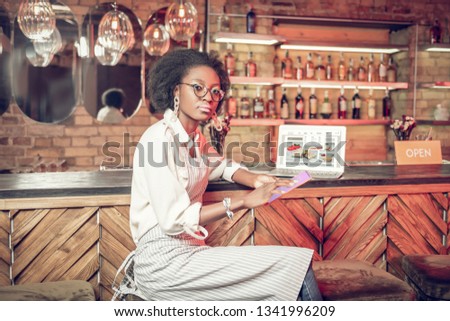 Clicking on table at bar. Serious pretty Afro-American curly-haired bar worker in glasses and white shirt with apron clicking on tablet while laptop is on bar counter