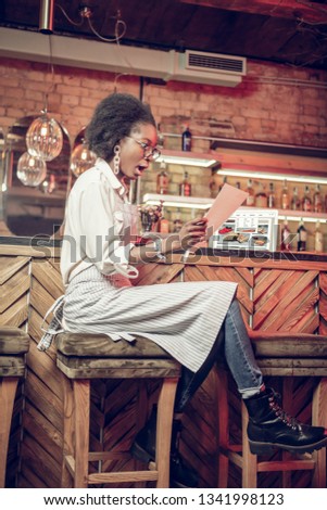 Woman with papers at bar. Surprised Afro-American dark-haired fancy bar employee with glasses wearing basic white shirt and dark skinny jeans looking at papers in hands in shock