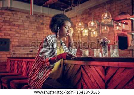 Considering menu at bar. Stylish sophisticated curly-haired Afro-American young female wearing striped cardigan sitting with coffee and looking at menu at bar
