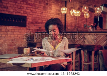 Fashion designer work. Nice-looking african-american creative designer with massive earrings wearing stylish comfy clothes anxiously looking at drawings at cafe table Royalty-Free Stock Photo #1341998243