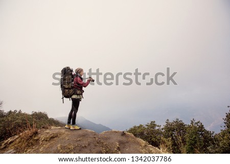Female traveller photographer with big backpack staying on the top of mountain in jungle forest. Take pictures and enjoy view in mountains trekking, Nepal 