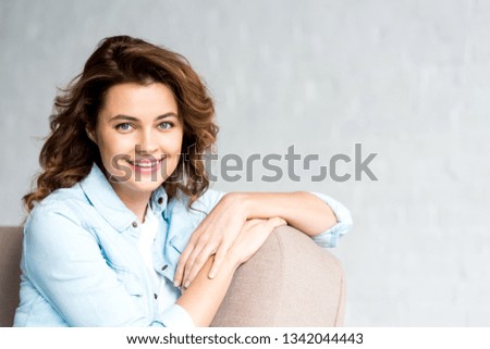 Happy brunette woman in shirt sitting on sofa and showing okay sign on grey 