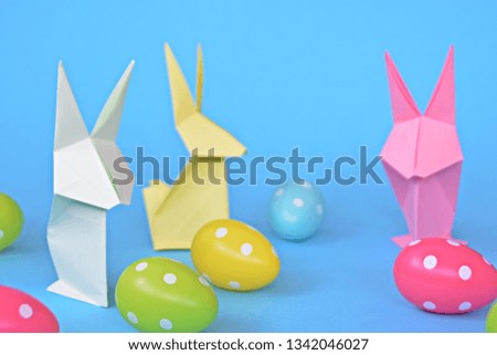 Origami bunnies folded from pastel paper stand on a blue base with dotted easter eggs on the outside - concept with origami for Easter with strong colors