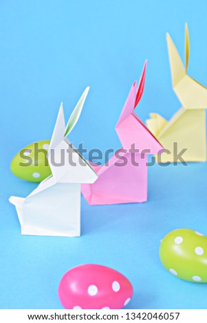 Origami bunnies folded from pastel paper stand on a blue base with dotted easter eggs on the outside - concept with origami for Easter with strong colors