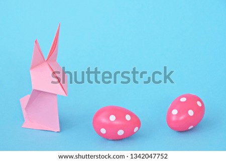 A pink origami bunny folded from pastel paper stands on a blue base on which are also Easter eggs with white dots - concept with origami and space for text or other elements for Easter