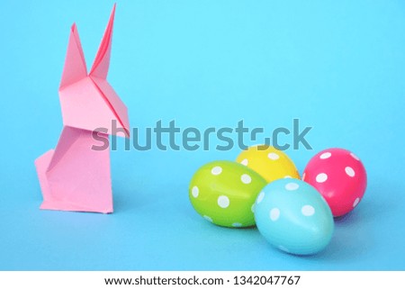 A pink origami bunny folded from pastel paper stands on a blue base on which are also Easter eggs with white dots - concept with origami and space for text or other elements for Easter