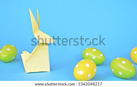 A yellow origami bunny folded from pastel paper stands on a blue base on which are also Easter eggs with white dots - concept with origami and space for text or other elements for Easter
