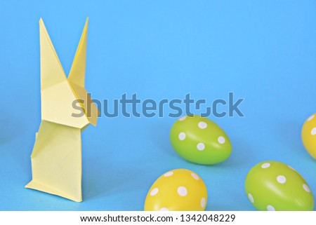 A yellow origami bunny folded from pastel paper stands on a blue base on which are also Easter eggs with white dots - concept with origami and space for text or other elements for Easter