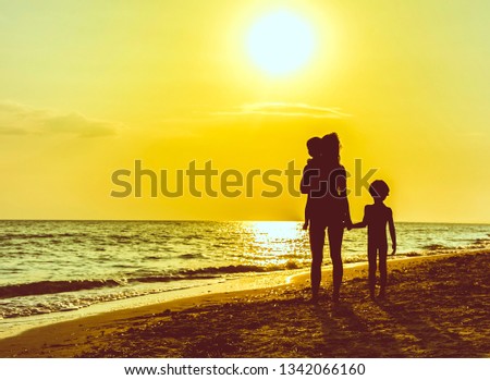 Silhouette of mother with two children on beach in sunset - Woman and child against direct sun ocean sunset - Parent holding and carrying children on beach in sunset looking away