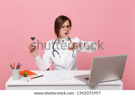 Female doctor sit at desk work on computer with medical document hold clock in hospital isolated on pastel pink wall background. Woman in medical gown glasses stethoscope. Healthcare medicine concept