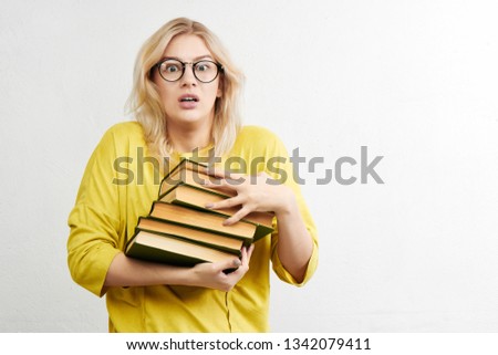 Frightened girl nerd blonde in round glasses with a worried face holding a stack of books on a white background in yellow clothes. Education and science concept
