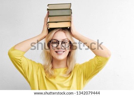 A beautiful woman in glasses is holding a stack of books above her head and is smiling toothyly on a white background. Happy and fun education