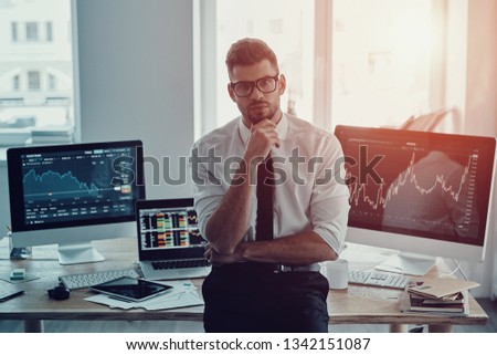 Modern businessman. Handsome young man in formalwear keeping hand on chin and looking at camera while standing in the office