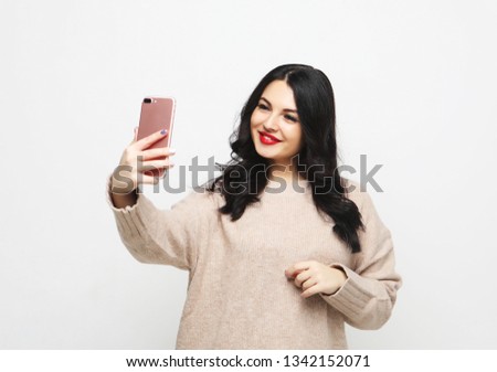 Fashion curvy brunette girl taking photo makes self portrait on smartphone wearing beige clothes over white  background