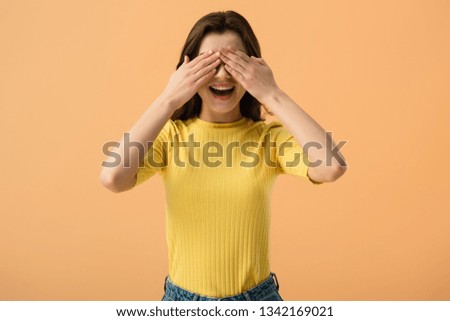 cheerful young woman covering eyes with hands isolated on orange 