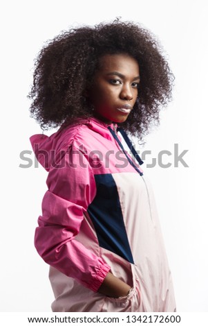 Saucy young curly brown-haired girl dressed in the pink sport jacket stands at the white background in the studio
